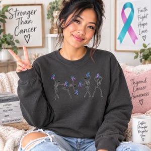May include: A dark charcoal gray crewneck sweatshirt featuring a graphic print of four white skeletons in various dancing poses, each holding a blue and pink awareness ribbon. The design is centered on the chest of the long-sleeved garment.