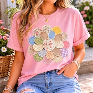 Puede incluir: Camiseta rosa claro con un gran diseño de flores aplicado. La flor está hecha de varios retazos de tela con estampados florales, a rayas y lisos. La camiseta tiene mangas cortas y un corte holgado.