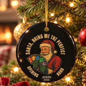 Op de afbeelding: Een rond zwart keramisch ornament met een illustratie van de Kerstman die vijf speelkaarten met elementaire symbolen vasthoudt. De tekst luidt: SANTA, BRING ME THE PERFECT OPENING HAND, JOHN, 2026. Het hangt aan een gouden koord.