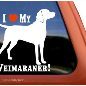 Op de afbeelding: Witte vinyl sticker met een silhouet van een Weimaraner hond met de tekst "I ❤️ MY WEIMARANER!". Het hart is oranje. De sticker is ontworpen om op een autoruit of een vergelijkbaar oppervlak te worden aangebracht.