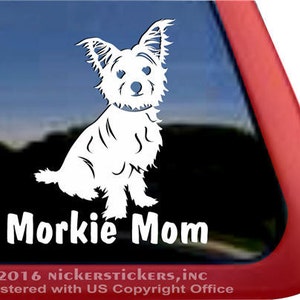 Könnte beinhalten: Weißer Vinyl-Aufkleber mit einem sitzenden Morkie-Hund und dem Text "Morkie Mom" darunter. Der Aufkleber befindet sich auf einem Fenster mit rotem Rand. Copyright-Informationen von Nickerstickers, Inc. sind unten sichtbar.