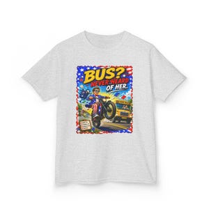 Puede incluir: Una camiseta gris claro con un estampado gráfico de una persona en una motocicleta y un autobús escolar amarillo al fondo. El texto dice BUS? NEVER HEARD OF HER. El diseño incluye detalles de estrellas en rojo, blanco y azul.