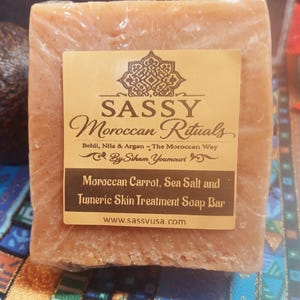 May include: A rectangular bar of soap wrapped in clear plastic. The soap has a light brown color and a label with the text "SASSY Moroccan Rituals" and "Moroccan Carrot, Sea Salt and Tumeric Skin Treatment Soap Bar."