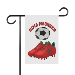 Puede incluir: Una bandera de jardín blanca con un balón de fútbol y botas de fútbol rojas con detalles verdes. El texto "DIMA MAGHREB" está arqueado sobre el balón en rojo. La bandera está montada en un soporte de metal negro.
