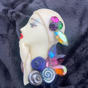 May include: A vintage-style brooch featuring a woman's profile in ivory with red lips and colorful gemstone accents. The brooch has a cluster of blue, silver, and iridescent embellishments, and is set against a dark, textured background.