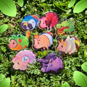 May include: A collection of eight colorful acrylic charms featuring stylized animal characters, including a blue creature, a pink pig with a rose, a green dragon, a frog, a pig with a crown, a bee-themed animal, a pink sheep, and a purple bull.
