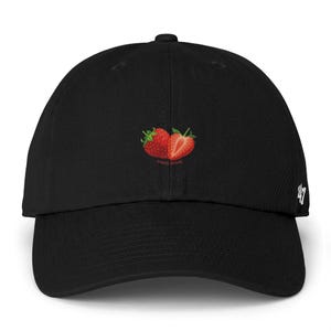 Puede incluir: Gorra de béisbol negra con un diseño bordado de dos fresas, una entera y otra cortada, en tonos rojos. La gorra tiene una visera curva y el número "47" es visible en el lateral.
