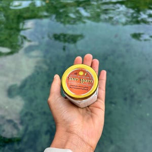 Könnte beinhalten: Ein kleines, sechseckiges Glas mit goldenem Deckel, beschriftet mit "sun yum" in roter und weißer Schrift. Das Glas wird in einer Hand vor dem Hintergrund von grünem Wasser gehalten. Der Text enthält auch die Hashtags #meetjade #remedible #sunscreennotfood.