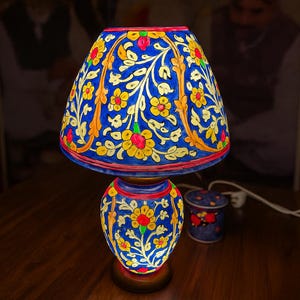 May include: A decorative table lamp with a blue base and shade, adorned with a floral pattern in yellow, red, and orange. The lamp is lit, casting a warm glow. A small matching container sits nearby.
