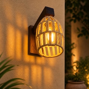 Puede incluir: Lámpara de pared para exteriores con pantalla de cuerda trenzada beige. La lámpara tiene una base y soporte de madera marrón oscuro y emite una luz cálida. La lámpara está montada en una pared clara y texturizada.