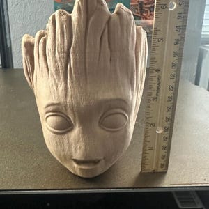 May include: A tan-colored, 3D-printed Groot-shaped planter, approximately 6 inches tall. The planter features a detailed face with large eyes and a small smile, and a textured, bark-like surface. The background includes toy cars.