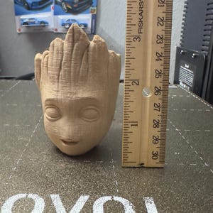 May include: A light brown, 3D-printed Groot planter, approximately 4 inches tall, with a detailed face and textured surface. A wooden ruler is placed next to the planter for scale. The background includes toy cars.
