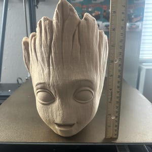 May include: A light brown, 3D-printed planter shaped like the character Groot from Guardians of the Galaxy. The planter is approximately 6 inches tall and features detailed facial features and textured bark-like surfaces. The planter is sitting on a black surface.
