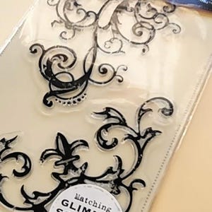 May include: Clear stamp set with black decorative flourish designs. The package includes the text "SCREEN PRINT CLEAR STAMPS" and a circular sticker that reads "Matching GLIMMER SCREENS available".