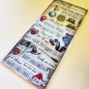 May include: A clear sheet of transparent stamps with various holiday themes, including Happy Birthday, Happy Valentine's Day, Happy 4th of July, Happy Easter, Happy Halloween, and Merry Christmas. The stamps are arranged in rows and columns, with each stamp featuring a different design. The sheet is packaged in a clear plastic sleeve.