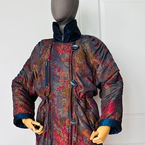 Peut inclure: Un long manteau matelassé doté d'une base gris foncé avec un motif géométrique multicolore dans des tons de rouge, orange et bleu. Le manteau comprend un col montant, une poche poitrine verticale zippée et une fermeture avant à trois boutons.