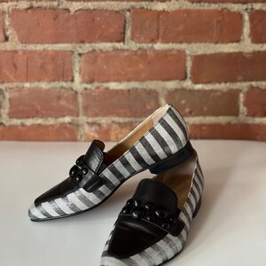 Peut inclure: Une paire de mocassins &agrave; rayures noires et blanches avec des d&eacute;tails en cuir noir. Les chaussures ont un ornement noir d&eacute;coratif sur le dessus et une semelle noire. Pr&eacute;sent&eacute;es sur une surface blanche avec un fond en briques.