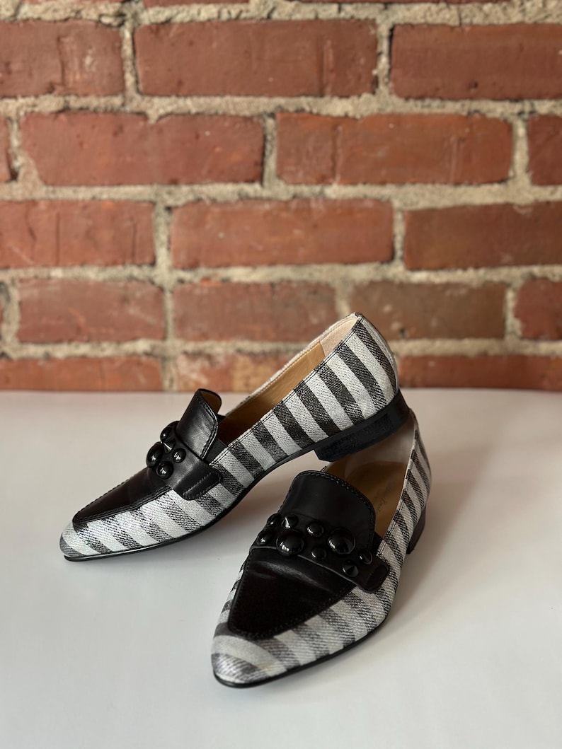 Peut inclure: Une paire de mocassins &agrave; rayures noires et blanches avec des d&eacute;tails en cuir noir. Les chaussures ont un ornement noir d&eacute;coratif sur le dessus et une semelle noire. Pr&eacute;sent&eacute;es sur une surface blanche avec un fond en briques.