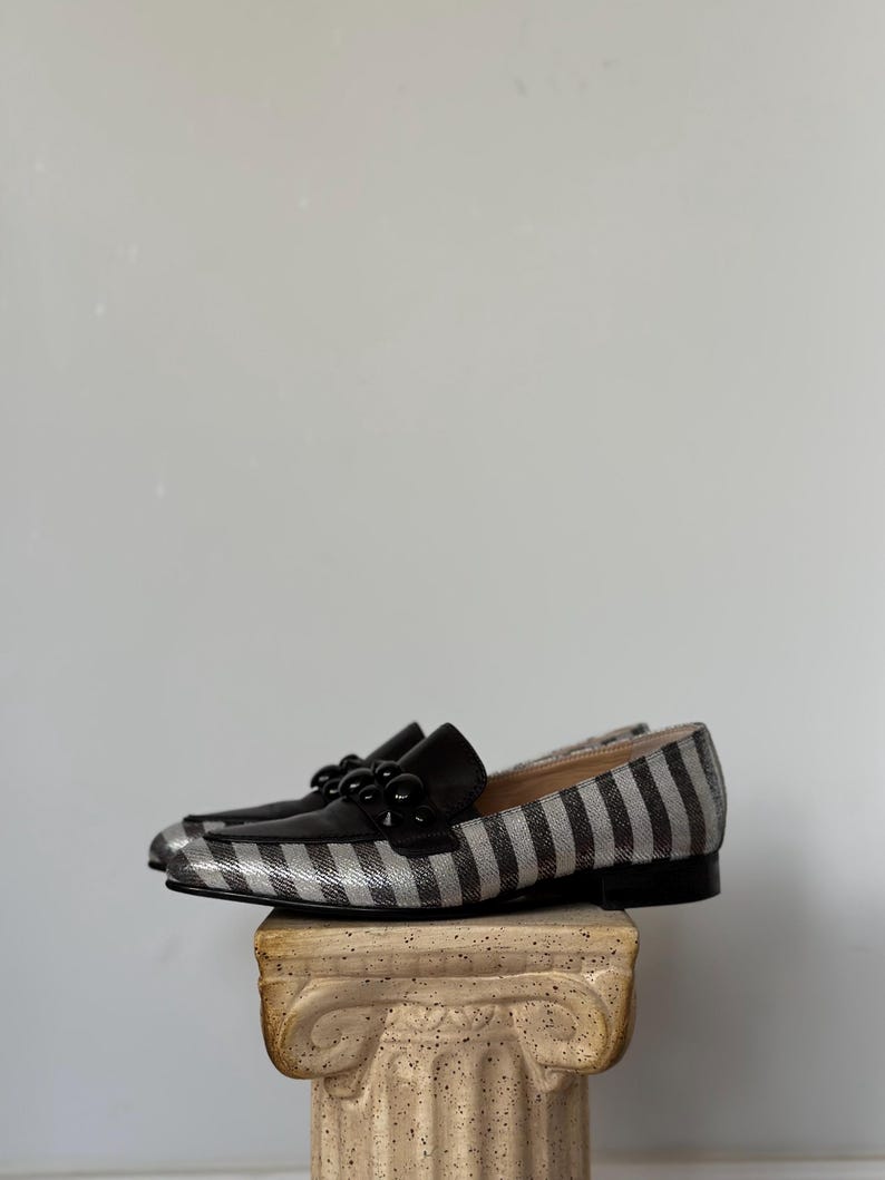 Peut inclure: Une paire de mocassins &agrave; rayures noires et argent&eacute;es avec des d&eacute;tails en cuir noir. Les chaussures ont une d&eacute;coration de perles noires sur le dessus et sont pr&eacute;sent&eacute;es sur un pi&eacute;destal en pierre.