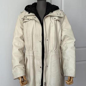 Op de afbeelding: Crèmekleurige jas met capuchon en zwarte binnenvoering. De jas heeft een ritssluiting, meerdere zakken en een taille met trekkoord. De jas wordt op een mannequin gepresenteerd.