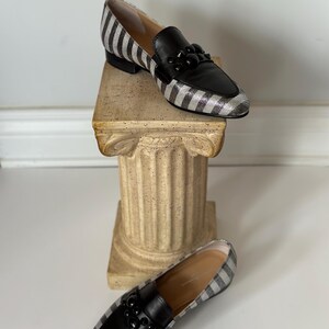 Peut inclure: Une paire de mocassins pour femmes avec un motif &agrave; rayures noires et argent&eacute;es. Les chaussures ont des empi&egrave;cements en cuir noir et une d&eacute;coration de perles noires. Les mocassins sont pr&eacute;sent&eacute;s sur une colonne d&eacute;corative.