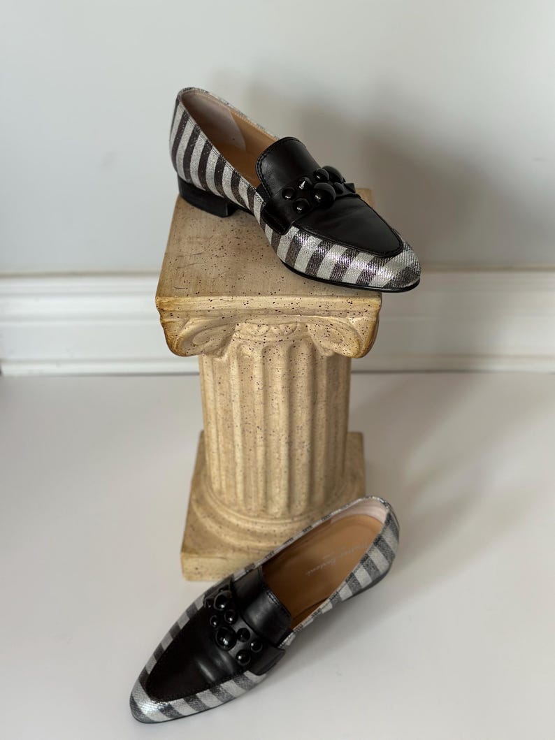 Peut inclure: Une paire de mocassins pour femmes avec un motif &agrave; rayures noires et argent&eacute;es. Les chaussures ont des empi&egrave;cements en cuir noir et une d&eacute;coration de perles noires. Les mocassins sont pr&eacute;sent&eacute;s sur une colonne d&eacute;corative.