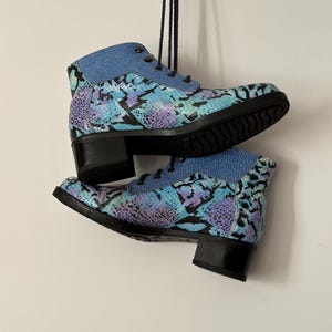 May include: Ankle boots with a snakeskin pattern in blue, purple, and black. The boots have a light blue collar and laces, with a black heel and sole. The boots are suspended against a white wall. The boots are approximately 15 cm high.