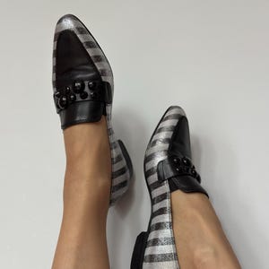 Peut inclure: Paire de mocassins ray&eacute;s argent et noir avec des d&eacute;tails en cuir noir. Les chaussures ont un bout arrondi et un petit talon noir. Une bride noire orn&eacute;e de perles noires orne le dessus de chaque chaussure.