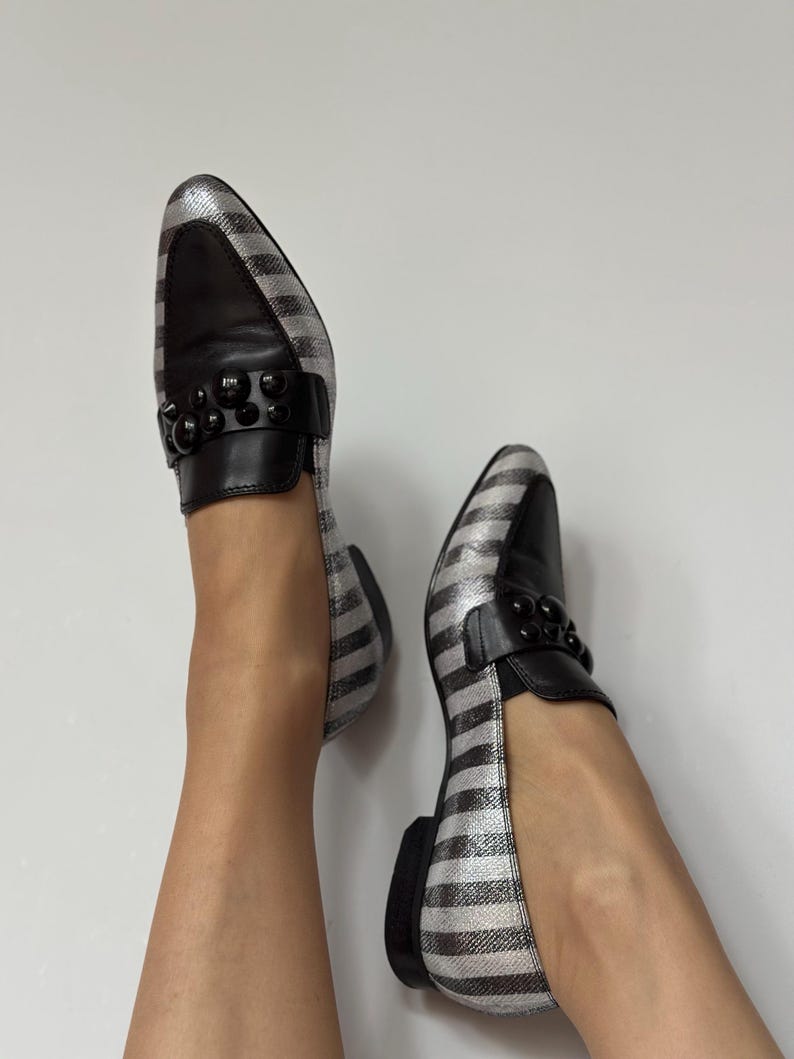 Peut inclure: Paire de mocassins ray&eacute;s argent et noir avec des d&eacute;tails en cuir noir. Les chaussures ont un bout arrondi et un petit talon noir. Une bride noire orn&eacute;e de perles noires orne le dessus de chaque chaussure.