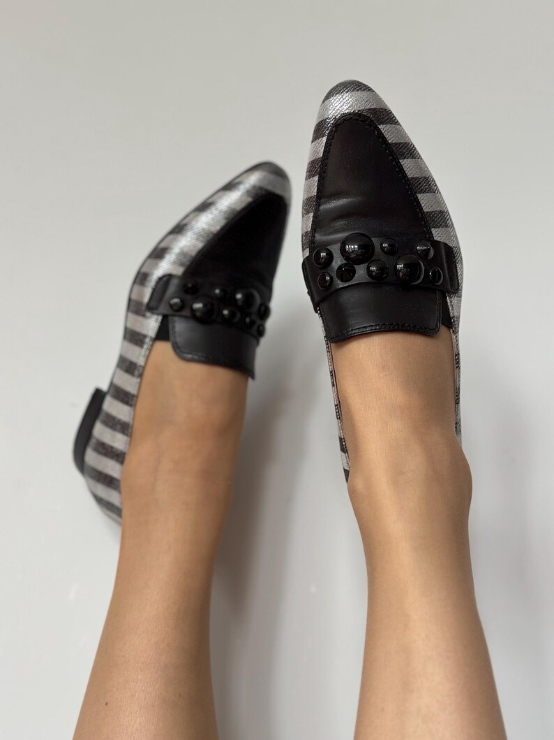 Peut inclure: Paire de mocassins &agrave; carreaux argent et noir avec des d&eacute;tails en cuir noir. Les chaussures ont un bout arrondi et une bride sur le dessus orn&eacute;e d'ornements sph&eacute;riques noirs. Fond blanc.