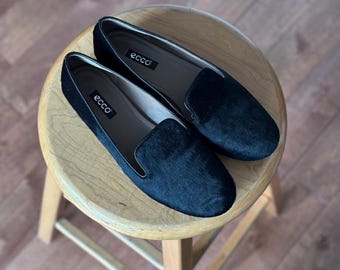 May include: A pair of black slip-on loafers with the brand name "ecco" visible on the insole. The shoes are made of a textured material and are resting on a small wooden stool.