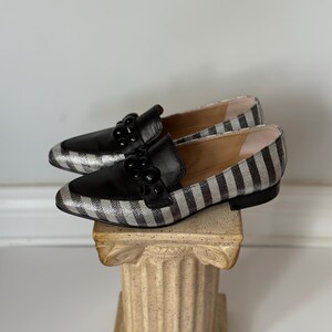 Peut inclure: Une paire de mocassins ray&eacute;s noirs et argent&eacute;s avec des d&eacute;tails en cuir noir. Les chaussures ont un bout pointu et sont orn&eacute;es d'ornements noirs. Les mocassins sont pr&eacute;sent&eacute;s sur une colonne d&eacute;corative.