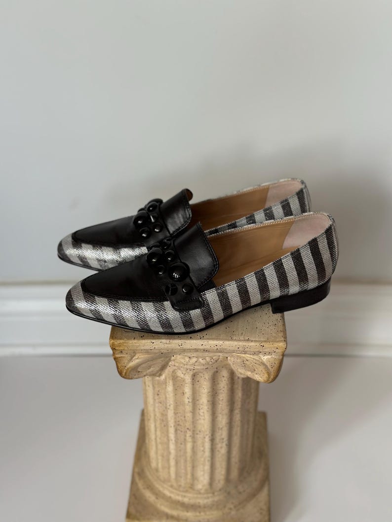 Peut inclure: Une paire de mocassins ray&eacute;s noirs et argent&eacute;s avec des d&eacute;tails en cuir noir. Les chaussures ont un bout pointu et sont orn&eacute;es d'ornements noirs. Les mocassins sont pr&eacute;sent&eacute;s sur une colonne d&eacute;corative.