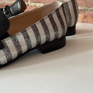 Peut inclure: Gros plan sur une paire de mocassins &agrave; rayures noires et blanches. Les chaussures ont une bride en cuir noir et un petit talon. L'int&eacute;rieur de la chaussure est de couleur beige clair. Le fond est un mur de briques et une surface blanche.