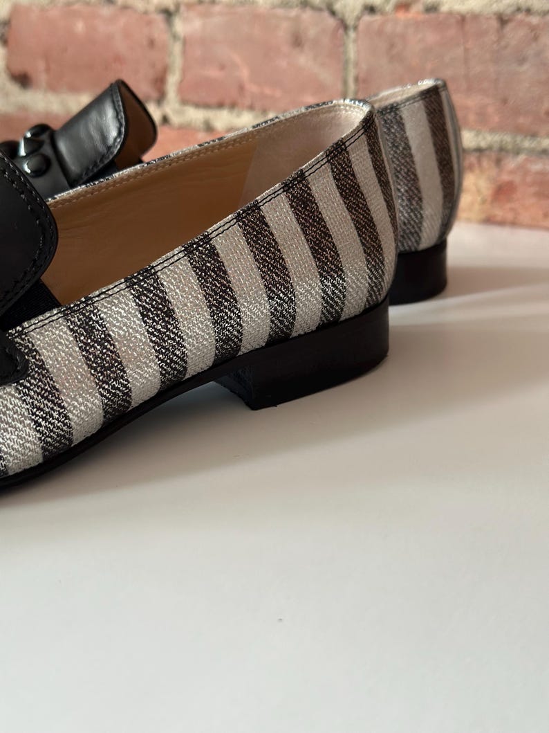 Peut inclure: Gros plan sur une paire de mocassins &agrave; rayures noires et blanches. Les chaussures ont une bride en cuir noir et un petit talon. L'int&eacute;rieur de la chaussure est de couleur beige clair. Le fond est un mur de briques et une surface blanche.
