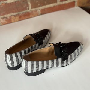 Peut inclure: Une paire de mocassins &agrave; rayures noires et blanches. Les chaussures ont un dessus en cuir noir et un petit talon. Le motif ray&eacute; est vertical et court sur les c&ocirc;t&eacute;s et le dessus. Les chaussures sont pos&eacute;es sur une surface blanche.
