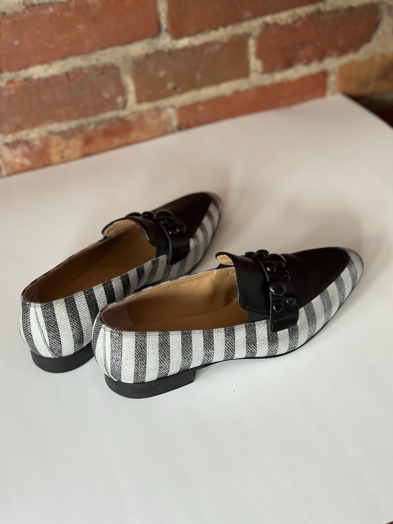 Peut inclure: Une paire de mocassins &agrave; rayures noires et blanches. Les chaussures ont un dessus en cuir noir et un petit talon. Le motif ray&eacute; est vertical et court sur les c&ocirc;t&eacute;s et le dessus. Les chaussures sont pos&eacute;es sur une surface blanche.