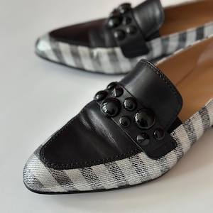 Peut inclure: Une paire de mocassins noirs et argent&eacute;s. Les chaussures ont une empeigne en cuir noir avec des clous noirs d&eacute;coratifs et un bout en tissu ray&eacute; argent&eacute; et blanc. Les chaussures sont mises au point sur un fond blanc.