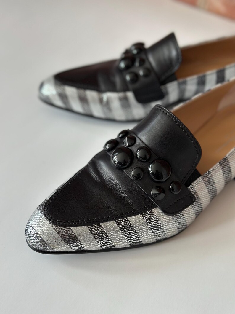 Peut inclure: Une paire de mocassins noirs et argent&eacute;s. Les chaussures ont une empeigne en cuir noir avec des clous noirs d&eacute;coratifs et un bout en tissu ray&eacute; argent&eacute; et blanc. Les chaussures sont mises au point sur un fond blanc.