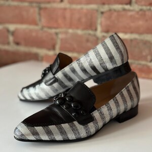 Peut inclure: Une paire de mocassins &agrave; rayures noires et blanches avec des d&eacute;tails en cuir noir. Les chaussures ont un design classique avec un bout arrondi et un petit talon. Le dessus de la chaussure comporte une bride en cuir noir avec des clous noirs d&eacute;coratifs.
