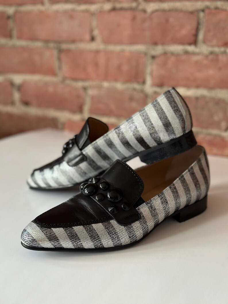 Peut inclure: Une paire de mocassins &agrave; rayures noires et blanches avec des d&eacute;tails en cuir noir. Les chaussures ont un design classique avec un bout arrondi et un petit talon. Le dessus de la chaussure comporte une bride en cuir noir avec des clous noirs d&eacute;coratifs.