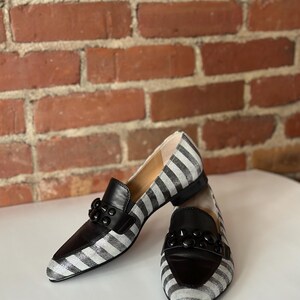 Peut inclure: Paire de mocassins pour femmes &agrave; rayures noires et blanches. Les chaussures ont des d&eacute;tails en cuir noir sur le bout et le cou-de-pied, et une d&eacute;coration noire. Les mocassins sont pos&eacute;s devant un mur de briques.
