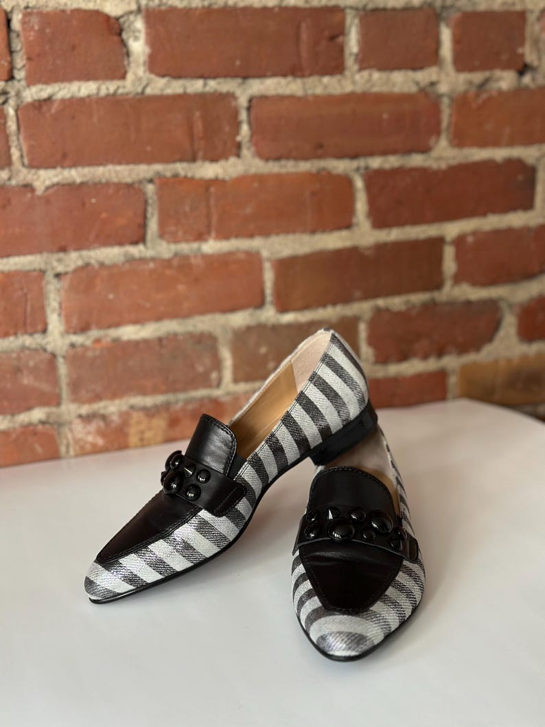 Peut inclure: Paire de mocassins pour femmes &agrave; rayures noires et blanches. Les chaussures ont des d&eacute;tails en cuir noir sur le bout et le cou-de-pied, et une d&eacute;coration noire. Les mocassins sont pos&eacute;s devant un mur de briques.