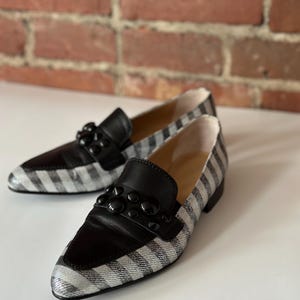 Peut inclure: Une paire de mocassins &agrave; rayures noires et blanches avec des d&eacute;tails en cuir noir. Les chaussures ont un bout pointu et sont orn&eacute;es d'ornements noirs sur le cou-de-pied. L'arri&egrave;re-plan est une surface blanche et un mur de briques.