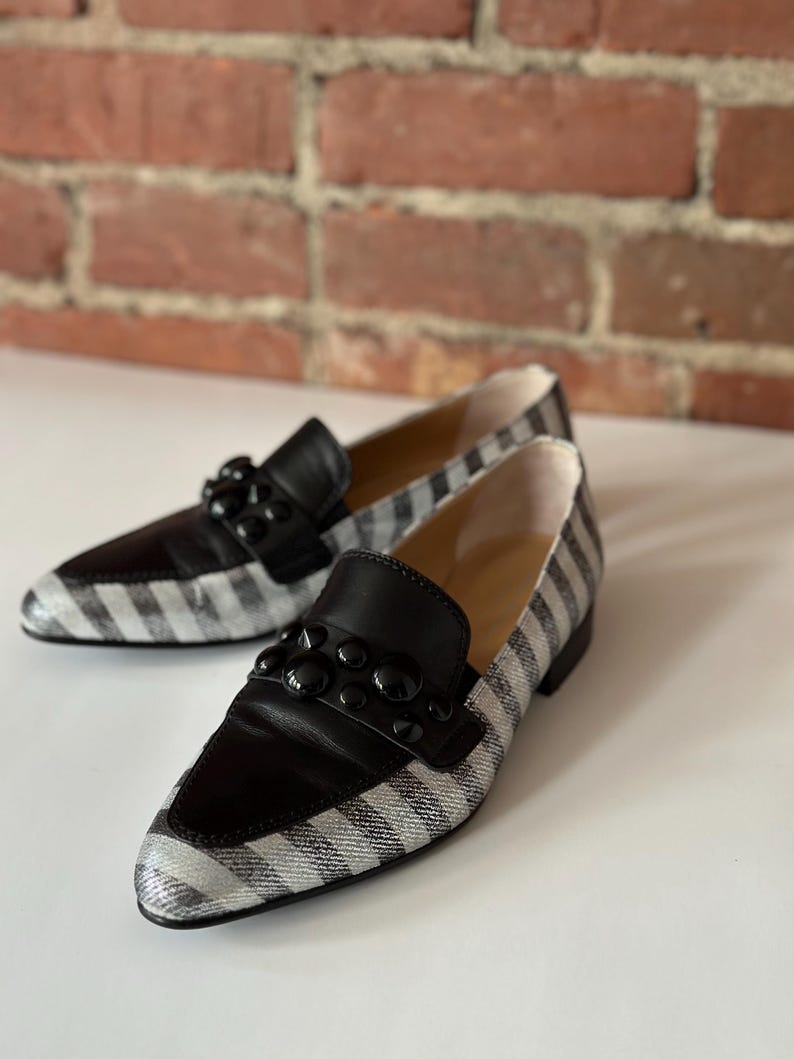 Peut inclure: Une paire de mocassins &agrave; rayures noires et blanches avec des d&eacute;tails en cuir noir. Les chaussures ont un bout pointu et sont orn&eacute;es d'ornements noirs sur le cou-de-pied. L'arri&egrave;re-plan est une surface blanche et un mur de briques.