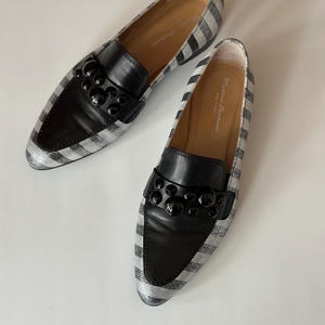 Peut inclure: Paire de mocassins ray&eacute;s noirs et argent&eacute;s avec des d&eacute;tails en cuir noir. Les chaussures ont un bout arrondi et une d&eacute;coration noire sur le cou-de-pied. L'int&eacute;rieur est de couleur beige clair.