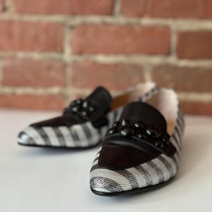 Peut inclure: Une paire de mocassins &agrave; rayures noires et blanches avec des d&eacute;tails en cuir noir. Les chaussures ont une d&eacute;coration de perles noires sur le dessus. Le fond est une surface blanche avec un mur de briques flou.