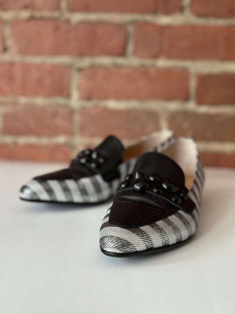 Peut inclure: Une paire de mocassins &agrave; rayures noires et blanches avec des d&eacute;tails en cuir noir. Les chaussures ont une d&eacute;coration de perles noires sur le dessus. Le fond est une surface blanche avec un mur de briques flou.