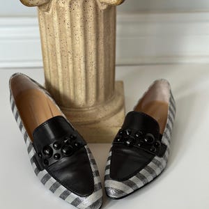 Peut inclure: Une paire de mocassins &agrave; rayures argent&eacute;es et noires avec des d&eacute;tails en cuir noir. Les chaussures ont un design classique avec un bout arrondi et un ornement noir d&eacute;coratif sur l'empeigne. Les chaussures sont pr&eacute;sent&eacute;es sur une surface blanche.