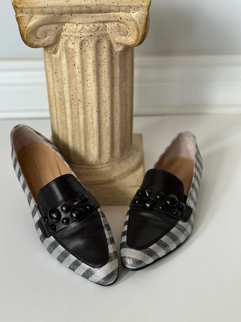 Peut inclure: Une paire de mocassins &agrave; rayures argent&eacute;es et noires avec des d&eacute;tails en cuir noir. Les chaussures ont un design classique avec un bout arrondi et un ornement noir d&eacute;coratif sur l'empeigne. Les chaussures sont pr&eacute;sent&eacute;es sur une surface blanche.